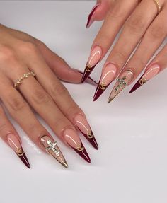 This striking design features Burgundy tips combined with gold embellishments on a nude base, creating a perfect contrast between boldness and elegance. The long stiletto shape of the nails accentuates the dramatic flair of the Burgundy tips, while the gold details, including tiny crescent moons and a cross, add a unique and luxurious touch. This look is ideal for someone who loves to mix classic colors with modern designs.   Photo credit by: @kayykrylics Maroon Stiletto Nails, Maroon Nail Ideas, Maroon Fall Nails, September Nails Art, Nails Maroon, Maroon Nail Designs, Maroon Nail, Burgundy Design, Old Money Nails