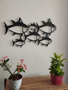 three metal fish on a white wall above a potted plant and some pink flowers