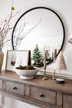 a wooden table topped with a bowl filled with pine cones and christmas trees in front of a round mirror