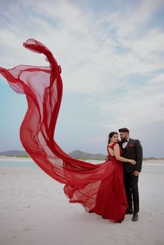 a man and woman standing on top of a beach next to each other holding a red scarf