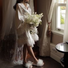 a woman standing in front of a window holding a bouquet of white flowers and feathers