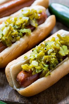 two hot dogs with toppings sitting on top of a cutting board next to pickles