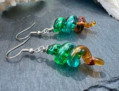 Enhance your accessory collection with these Unique Lampwork Glass Dangle Earrings featuring  hollow dangles in transparent emerald, teal, and topaz. Measuring 2" long, these stylish earrings are sure to make a statement wherever you go. Perfect for the fashion-forward individual who appreciates unique and handcrafted jewelry. Handmade by skilled glass artisan, Alisha White. Embrace the blend of artistic tradition and modern style with these handcrafted treasures, and let your earrings be a conv Green Glass Dangle Jewelry, Green Teardrop Glass Jewelry, Green Earrings For Jewelry Making, Green Drop Earrings With Unique Variations, Modern Green Glass Earrings, Hypoallergenic Green Glass Jewelry, Green Dangle Glass Earrings, Green Glass Dangle Earrings, Nickel-free Green Glass Earrings