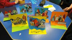 children are sitting at a table making cards with paper houses and other things on them