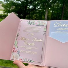 a person holding up a pink and gold wedding program book in front of some trees