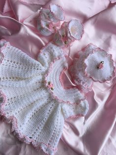 a crocheted baby dress and bonnet laying on a pink satin bed sheet with flowers