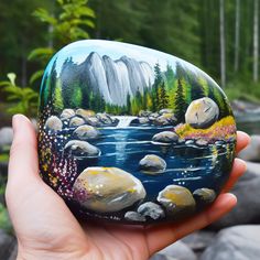 a hand holding a painted rock in front of a river and mountain scene with pine trees