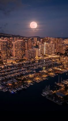 the full moon is seen over a city at night with boats docked in the harbor