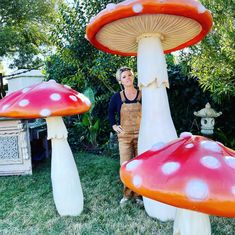 a woman standing in front of two giant mushroom shaped structures on the grass with trees and bushes behind her