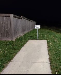 a white sign sitting on the side of a sidewalk next to a wooden fence at night