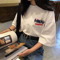 a woman sitting on the floor holding a magazine and looking at her cell phone while wearing a white t - shirt