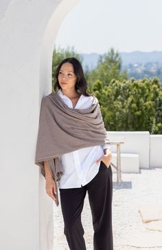 a woman standing next to a white wall wearing a gray shawl and black pants