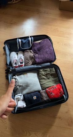 an open suitcase filled with clothes on top of a hard wood floor next to a person's hand