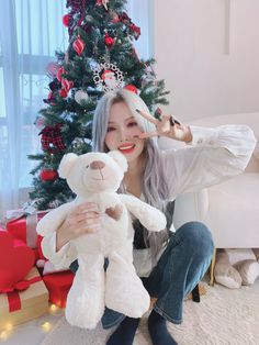 a woman sitting in front of a christmas tree holding a teddy bear and giving the peace sign