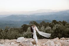 a bride and groom standing on top of a rocky hill with mountains in the background