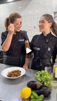 two women in black aprons standing next to a counter with plates of food on it