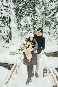 a man and woman are standing on a bridge in the snow holding bouquets with flowers