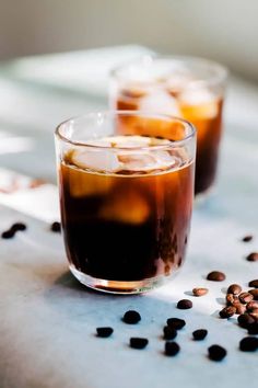 two glasses filled with iced coffee sitting on top of a table next to some beans