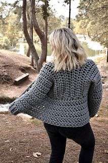 a woman standing in the woods with her back to the camera, wearing a gray crocheted sweater