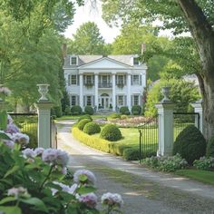 a large white house surrounded by lush green trees and flowers in front of it's gate