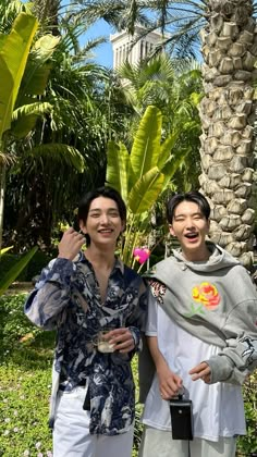 two young men standing next to each other in front of trees and flowers, one holding a cell phone