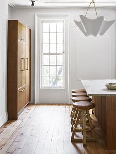 a kitchen with white walls and wooden flooring next to a large window, bar stools in front of the counter