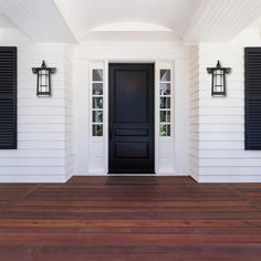 a black door is on the side of a white house with shutters and wood flooring