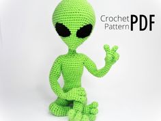 a crocheted alien sitting on the ground with its hands up and eyes closed