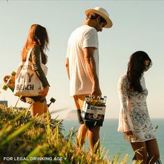 three people standing on the side of a hill looking at the ocean and holding bags