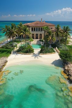 an aerial view of a large house with palm trees in the foreground and clear blue water on the other side