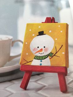 a small wooden easel with a painted snowman on it and a cup of coffee in the background