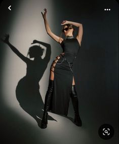 a woman in a black dress and knee high boots posing for a photo with her shadow on the wall