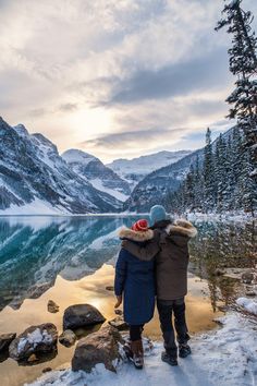 two people standing next to each other in front of a lake with snow on the ground