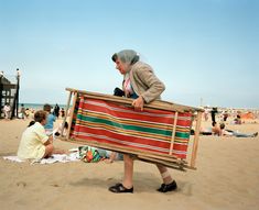 a woman carrying a bench on the beach with people sitting in the sand behind her