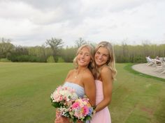 two beautiful young women standing next to each other on a lush green field with trees in the background