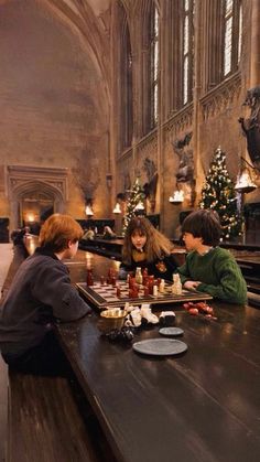 three children sitting at a long table playing chess in a large room with high ceilings