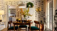 a dining room table surrounded by chairs in front of an open kitchen door with floral wallpaper on the walls