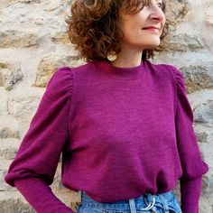 a woman with curly hair wearing a purple sweater and jeans standing in front of a stone wall