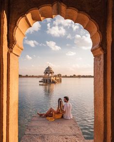 two people sitting on the edge of a body of water with an arch in front of them