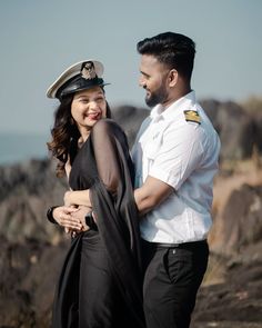 a man and woman standing next to each other on top of a rocky hill with the ocean in the background