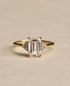 a gold ring with an emerald stone and three diamonds on the band, sitting on a beige surface