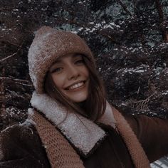 a woman wearing a hat and scarf standing in the snow with trees behind her looking off into the distance