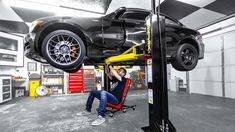 a man sitting in a chair under a car lifted by a lift with his feet on the ground