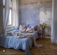 an unmade bed sitting in front of a window next to a table with flowers on it