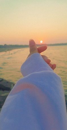 someone is holding their hand out to the water at sunset or sunrise, with the sun setting in the distance