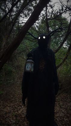 a person in a dark costume with glowing eyes and headlamps standing in the woods