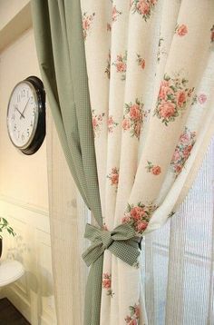 the clock is hanging on the wall next to the window with floral curtains in it