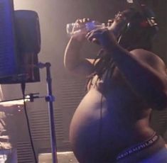 a pregnant woman drinking from a glass in front of a microphone and recording equipment behind her