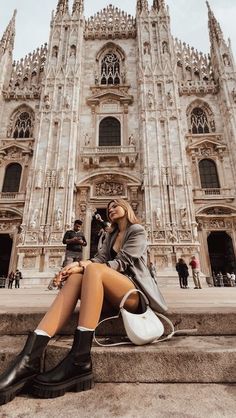 a woman sitting on the steps in front of a cathedral with her legs crossed and wearing black boots