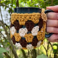 a hand holding a crocheted coffee mug in front of some trees and bushes